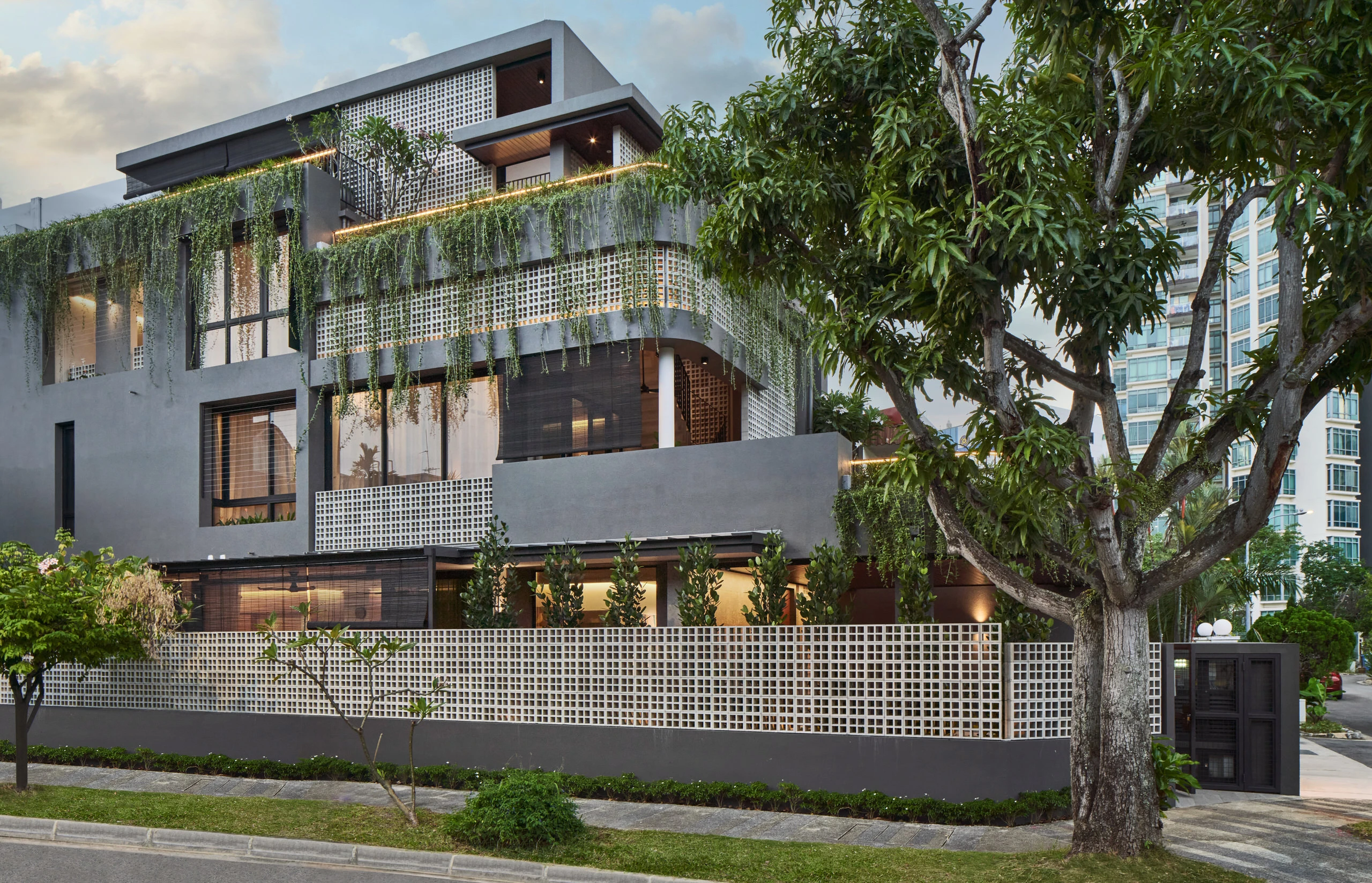 Three-storey tropical house with breeze-block screens, cascading greenery on balconies, and mature street trees at dusk