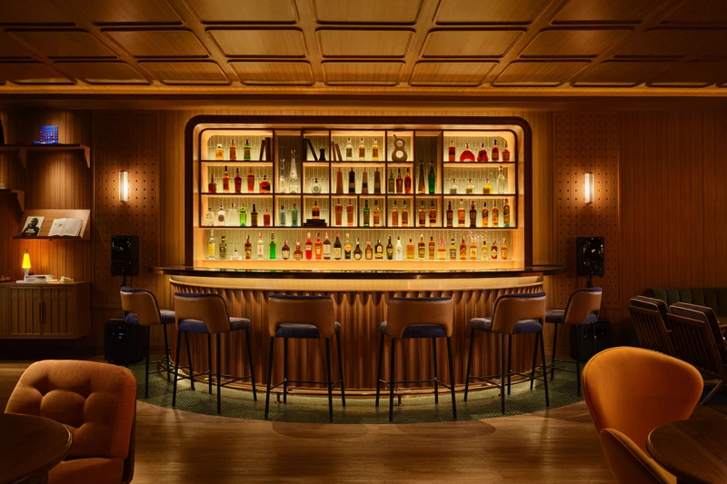 Curved wood-paneled bar with backlit bottle display, leather stools, and coffered ceiling in warm ambient lighting