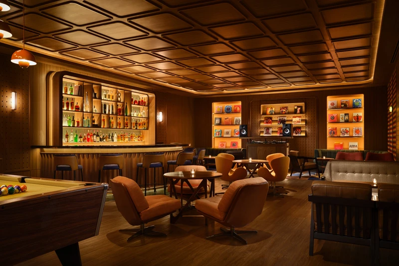 Retro lounge with wood paneling, backlit bar, pool table, swivel armchairs, and illuminated display shelves